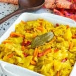 A white serving dish filled with Caribbean Steamed Cabbage garnished with a bay leaf, sits next to a dark wooden spoon and an orange kitchen towel.