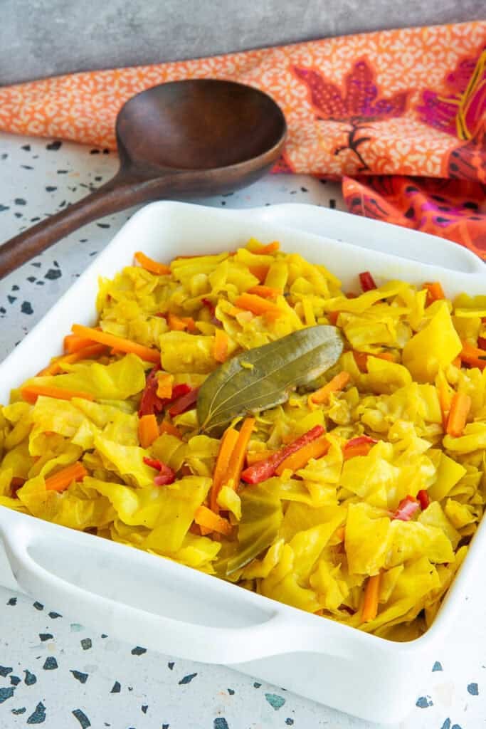 A white serving dish filled with Caribbean Steamed Cabbage garnished with a bay leaf, sits next to a dark wooden spoon and an orange kitchen towel.