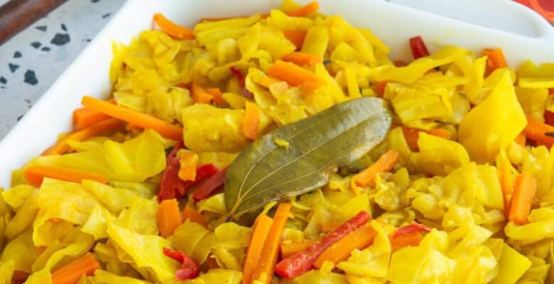 A white serving dish filled with Caribbean Steamed Cabbage garnished with a bay leaf, sits next to a dark wooden spoon and an orange kitchen towel.