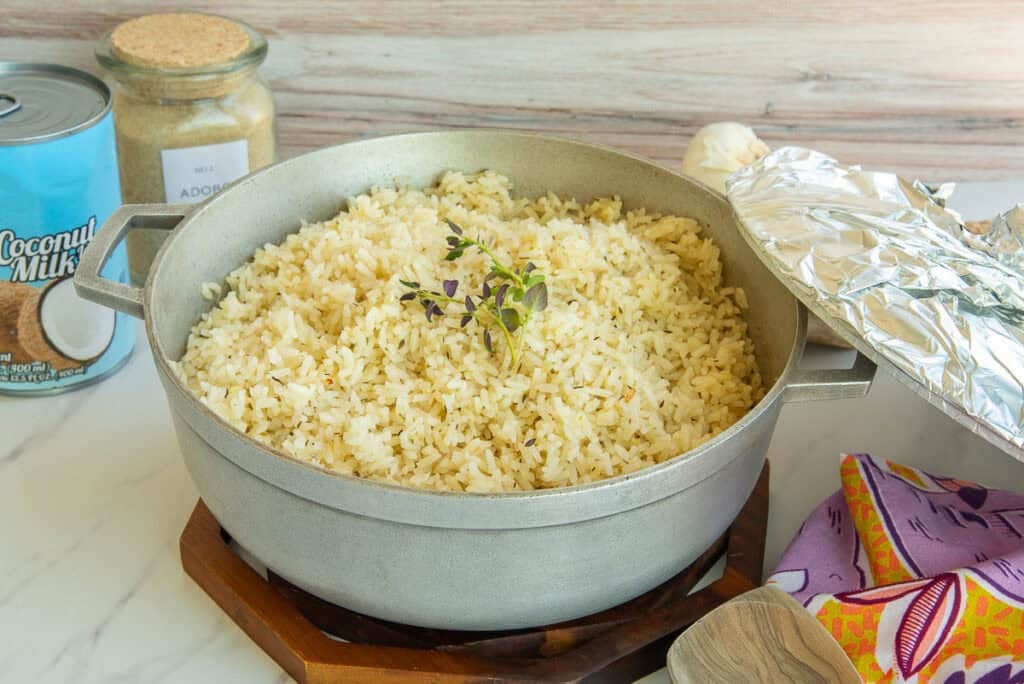 A pot of Caribbean Coconut Rice garnished with a sprig of thyme sits in front of a can of coconut milk and a jar of adobo seasoning blend.
