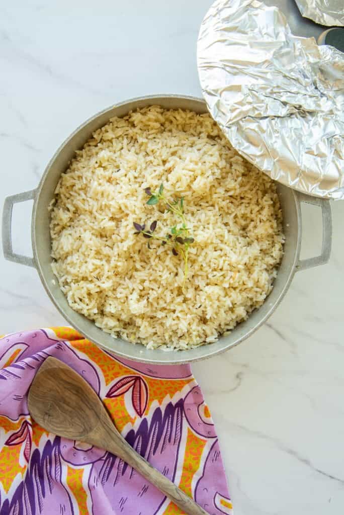 A pot of Caribbean Coconut Rice is next to a purple kitchen towel and a wooden spoon.