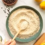 A spoon swirls the lemon pepper adobo seasoning in a dark green ceramic bowl that's surrounded by its ingredients.
