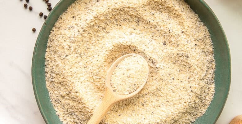 A spoon swirls the lemon pepper adobo seasoning in a dark green ceramic bowl that's surrounded by its ingredients.