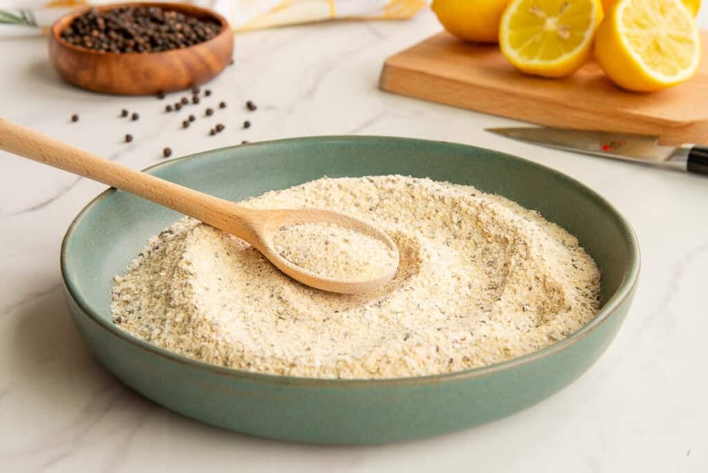 A spoon is swirled around in the Lemon Pepper Adobo Seasoning blend which is in a green ceramic bowl.