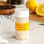 The lemon pepper adobo seasoning is bottled in a plastic spice jar that's sitting in front of a cut lemon and a bowl of peppercorns.
