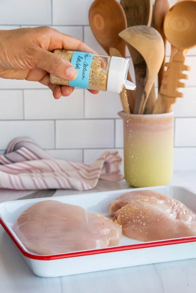 A hand shaking low-sodium adobo from a plastic bottle onto raw chicken breasts on a sheet pan.