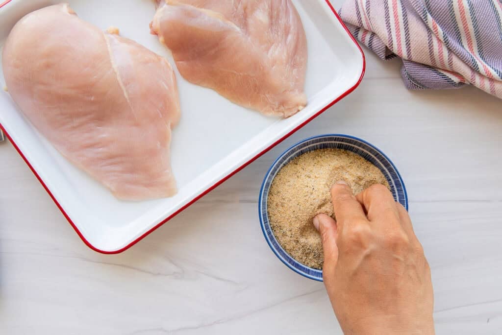 A hand pinches the adobo from a blue prep bowl that's next to a sheet pan with raw chicken breasts on it.
