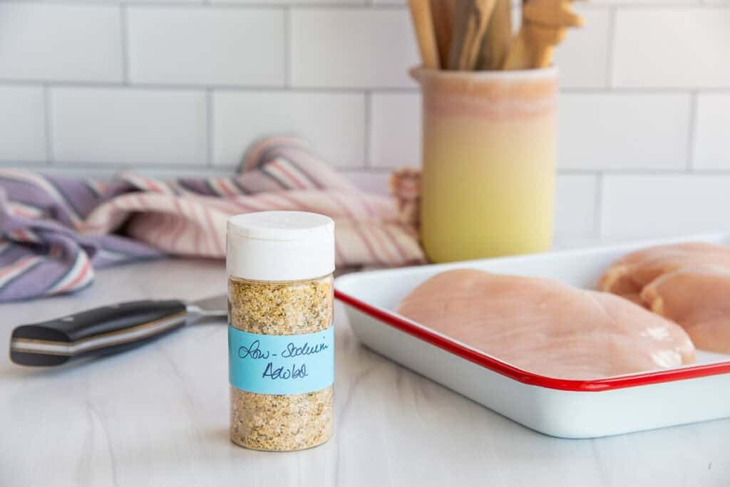 A bottle of low-sodium adobo sits on a white kitchen counter next to a pan with raw chicken breasts on it.