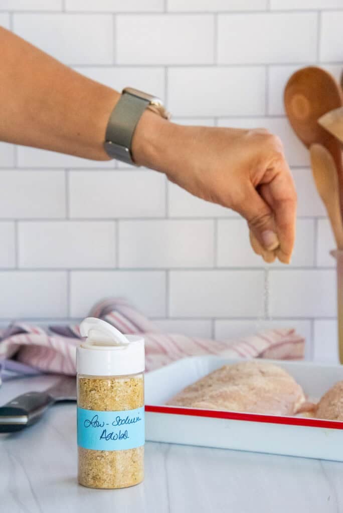 A bottle of low-sodium adobo sits in front of a sheet pan with chicken on it and a hand sprinkling the seasoning blend over the meat.