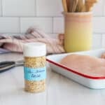 A small bottle of low-sodium adobo on a kitchen counter next to a pan holding two raw chicken breasts.