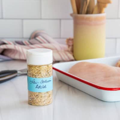 A small bottle of low-sodium adobo on a kitchen counter next to a pan holding two raw chicken breasts.