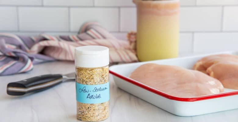 A small bottle of low-sodium adobo on a kitchen counter next to a pan holding two raw chicken breasts.