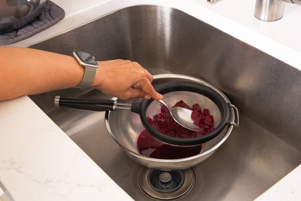 A wide, flat metal spoon presses the juice from the softened fruit through a fine-mesh sieve into a metal bowl that's sat in a sink.