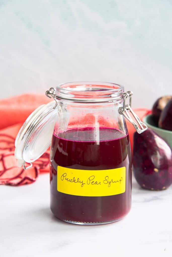 A glass jar holds prickly pear syrup and sits next to a prickly pear and a coral kitchen towel.