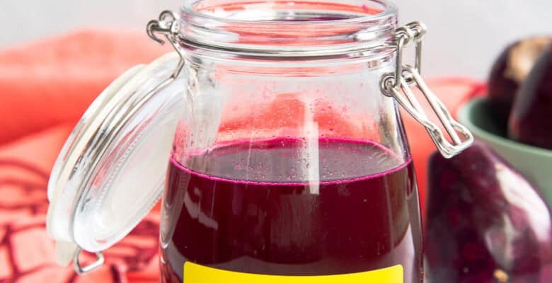 A glass jar holds prickly pear syrup and sits next to a prickly pear and a coral kitchen towel.