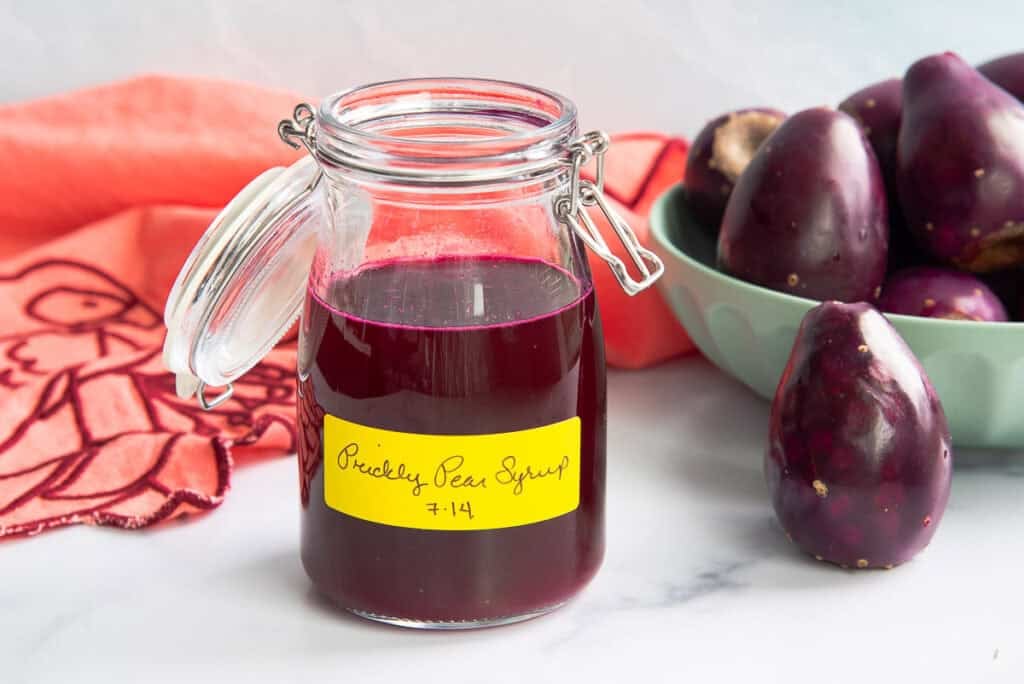 Prickly Pear Syrup in a glass jar with a swing-top lid and sits next to a green bowl filled with prickly pears.