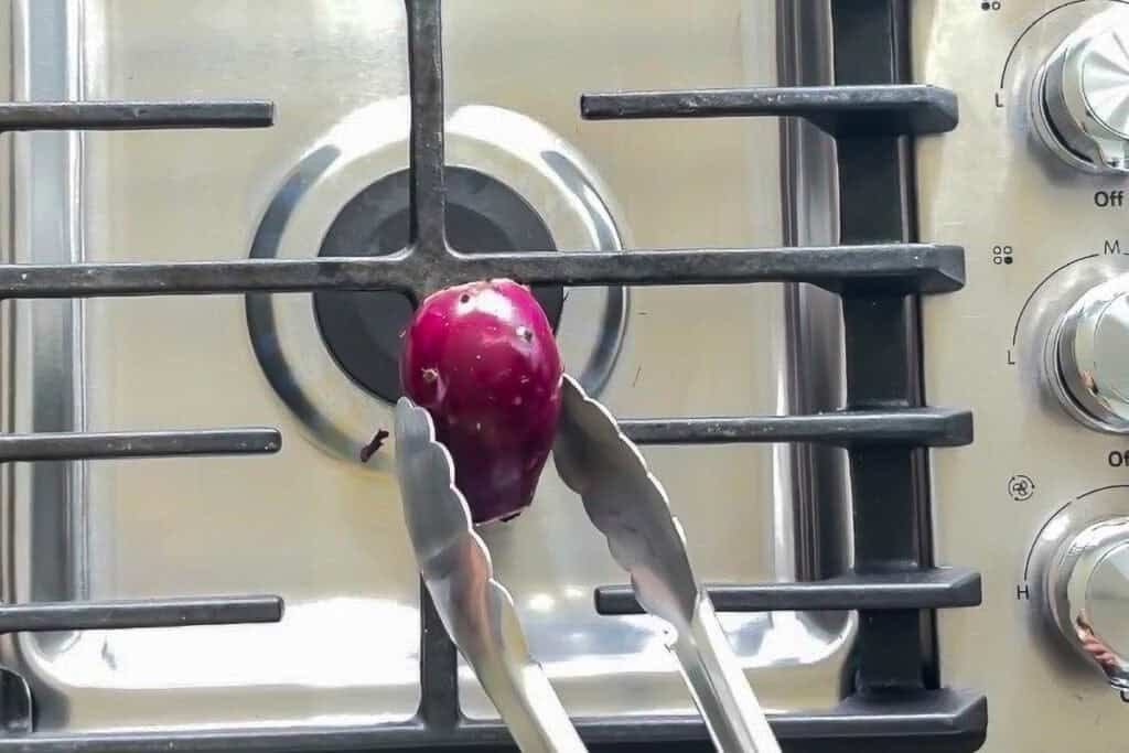 Tongs hold a prickly pear over the open flame of a gas stovetop.