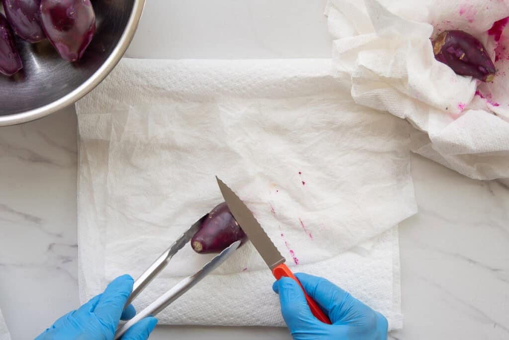 Marta's blue-gloved hands uses the back of a paring knife to scrape away the glochids on the fruit while holding it with a pair of tongs.