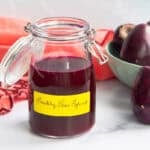 A glass jar with a swing-top lid filled with Prickly Pear Syrup sits in front of a bowl filled with prickly pears with one pear sitting next to the jar.