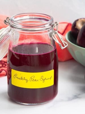 A glass jar with a swing-top lid filled with Prickly Pear Syrup sits in front of a bowl filled with prickly pears with one pear sitting next to the jar.