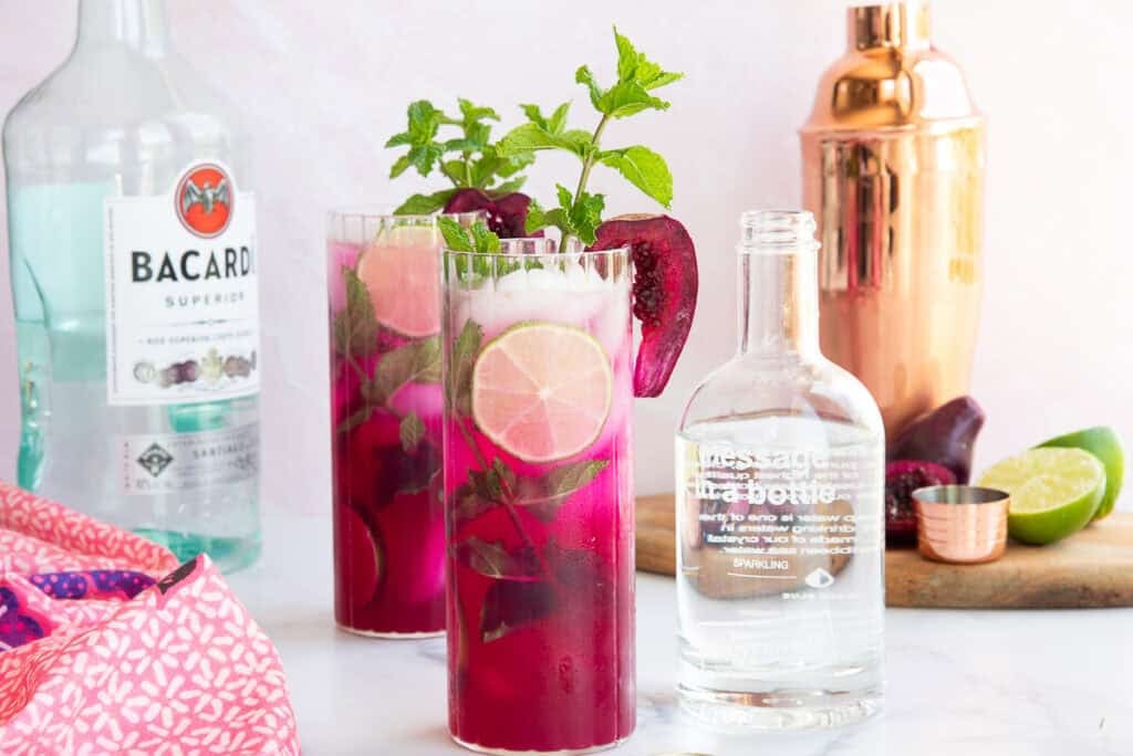 Two glasses filled with prickly pear mojito garnished with fresh prickly pear slices, mint and sitting next to a bottle of sparkling soda, white rum, and a copper cocktail shaker.