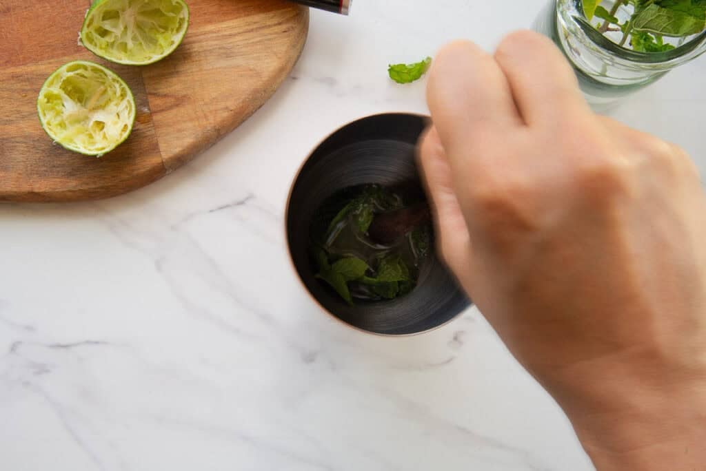 A muddler is used to bruise the mint leaves in a silver cocktail shaker.