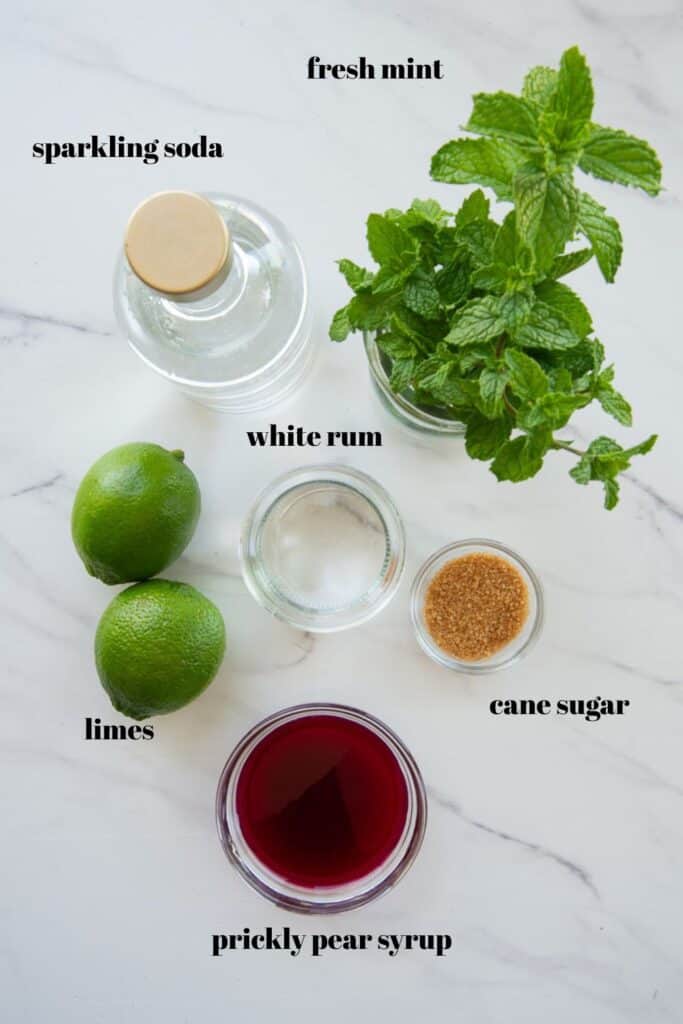 The ingredients needed to make Prickly Pear Mojitos are on a white countertop and labeled with black text.