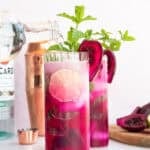 Sparkling water is poured from a glass bottle into a highball glass filled Prickly Pear Mojitos that's garnished with a slice of prickly pear fruit and fresh mint.