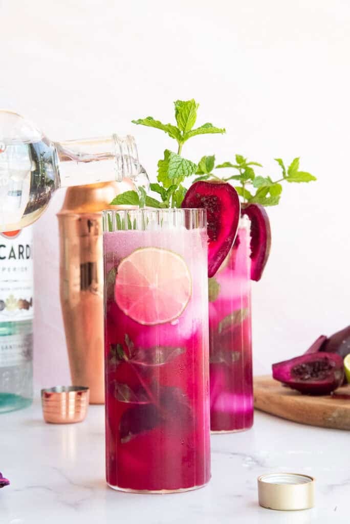 Sparkling water is poured from a glass bottle into a highball glass filled Prickly Pear Mojitos that's garnished with a slice of prickly pear fruit and fresh mint.