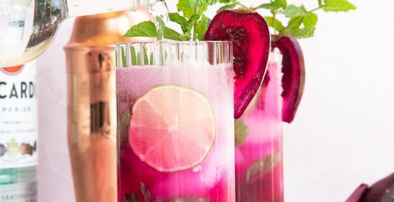Sparkling water is poured from a glass bottle into a highball glass filled Prickly Pear Mojitos that's garnished with a slice of prickly pear fruit and fresh mint.